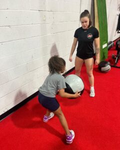 Young athlete in a gym performing a squat with a large medicine ball, supervised and trained by a Elite Edge Trainer for strength and conditioning.
